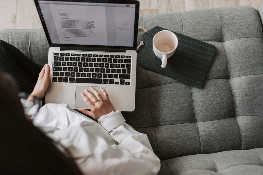 A person is sitting on a sofa, typing on a laptop on their lap. There is a cup of coffee on the sofa beside them.