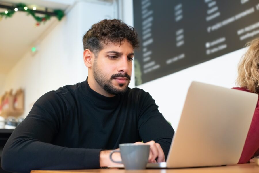 A man sitting in a coffee shop is looking at his laptop with a concerned look on his face. He has a cup of coffee beside him.