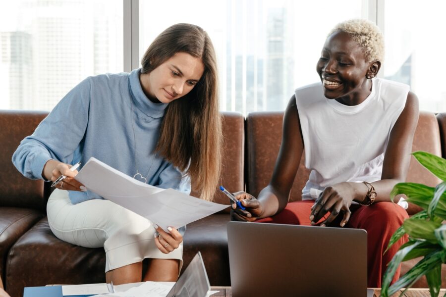 Two women sit on a couch looking at sheets of paper. There is a laptop in front of them on a low table.