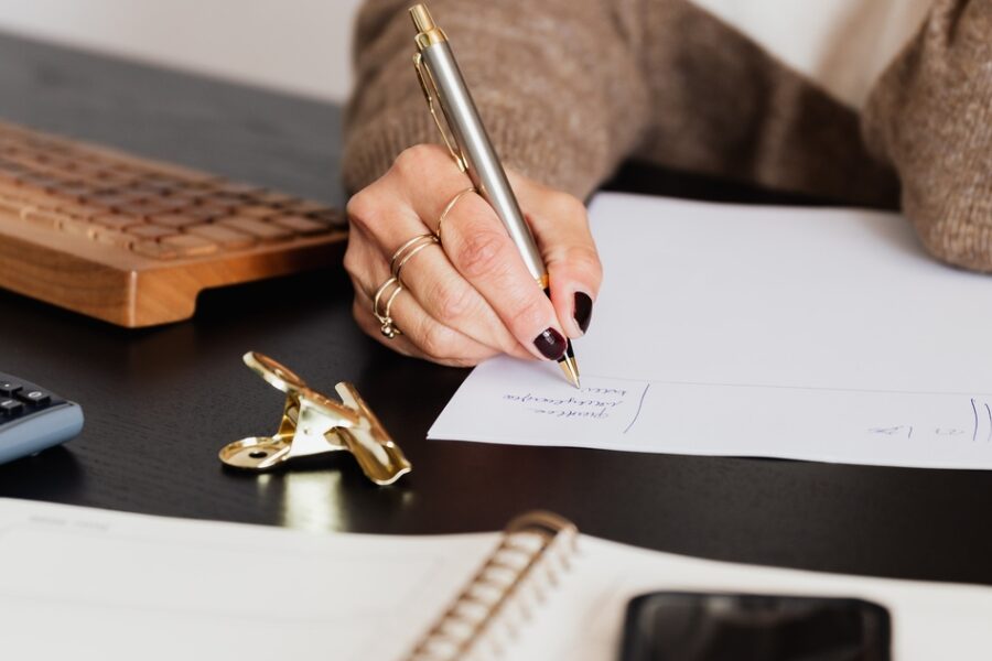 A person is writing on a sheet of paper on their desk. Beside them, there is a computer keyboard, and in front of them is a pad of paper, a phone, and other office supplies.