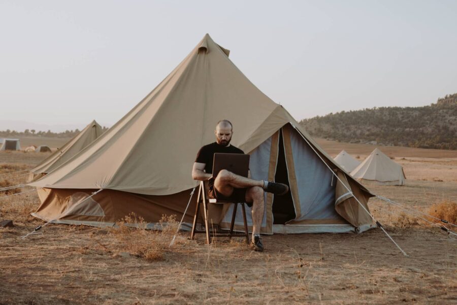 A man is sitting in a chair in front of a tent in the middle of a deserted area. He has a laptop on his lap.