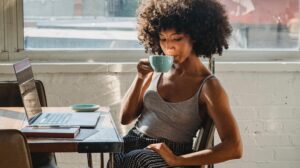 A woman is sitting at a desk drinking a cup of coffee. On the desk in front of her, there is a laptop and a notepad. 