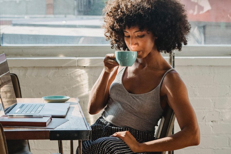 A woman is sitting at a desk drinking a cup of coffee. On the desk in front of her, there is a laptop and a notepad.