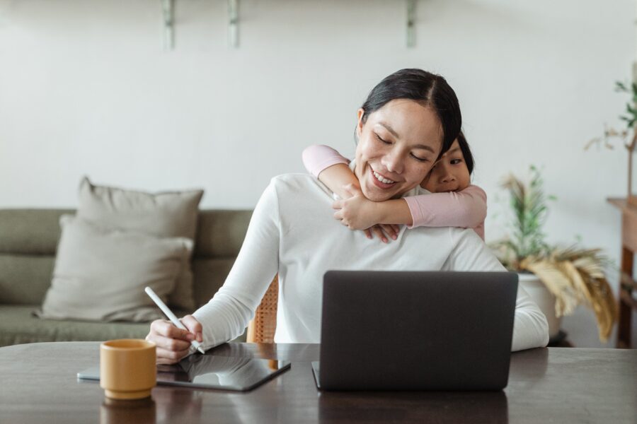 A dark-haired woman is sitting at a desk, looking at a laptop, and writing on a tablet with a stylus. Behind her, a young child is hugging the woman around her shoulders. The woman is smiling.