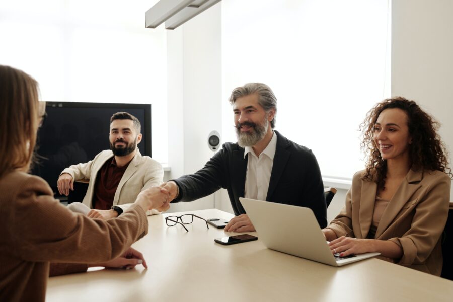 A group of workers are seated around a large desk in an office space. From one side of the table, a man with a grey beard is shaking the hand of another person seated across from him. He and the others seated at the table with him are smiling.