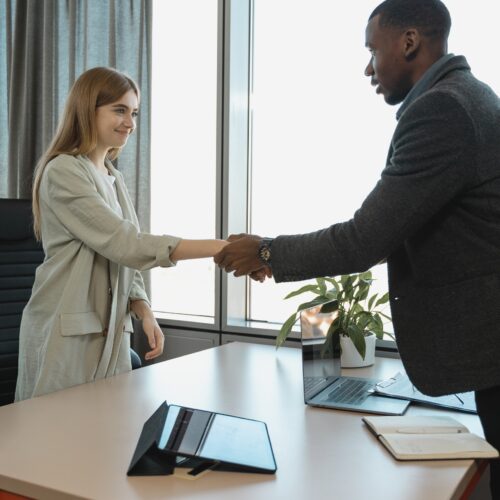 A Black man with very short, dark hair in a dark grey wool blazer and dress pants is shaking the hand of a white woman with strawberry blonde, long hair pushed behind her shoulders. She is wearing a mint-coloured, lightweight, long blazer. The woman is standing behind the man's desk. They are shaking hands while standing over the table, where there are office supplies, including a note pad, laptop, tablet, and a potted plant.