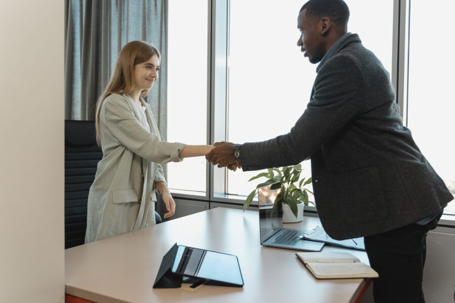 A Black man with very short, dark hair in a dark grey wool blazer and dress pants is shaking the hand of a white woman with strawberry blonde, long hair pushed behind her shoulders. She is wearing a mint-coloured, lightweight, long blazer. The woman is standing behind the man's desk. They are shaking hands while standing over the table, where there are office supplies, including a note pad, laptop, tablet, and a potted plant.