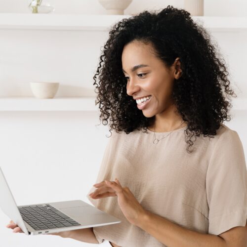 A Black woman with tight curly hair to her shoulders is smiling, looking at the laptop open in her right hand. She is scrolling with her left hand. She is wearing a neutral coloured shirt, which matches the neutral, contemporary office space behind her with plain shelves and a few cream coloured decorative bowls.