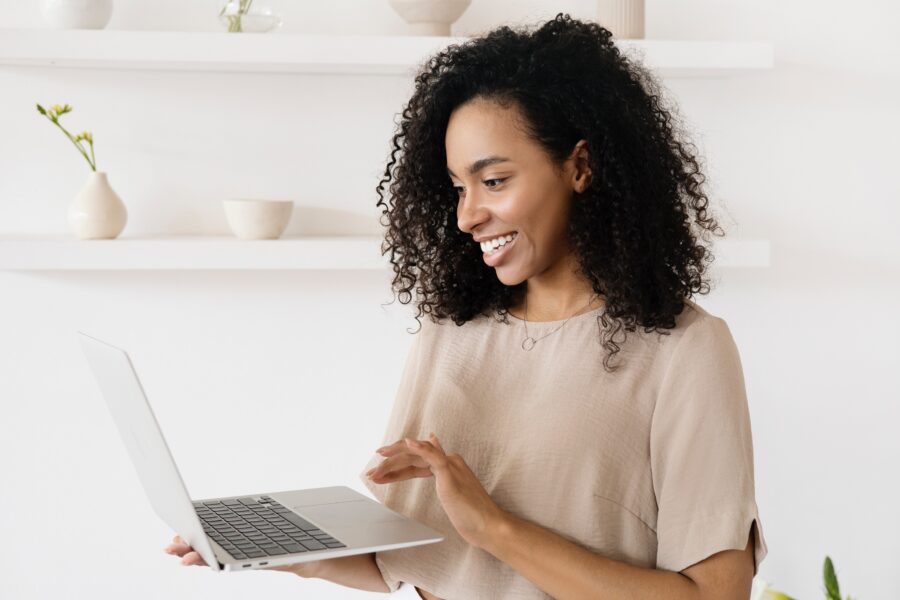 A Black woman with tight curly hair to her shoulders is smiling, looking at the laptop open in her right hand. She is scrolling with her left hand. She is wearing a neutral coloured shirt, which matches the neutral, contemporary office space behind her with plain shelves and a few cream coloured decorative bowls.