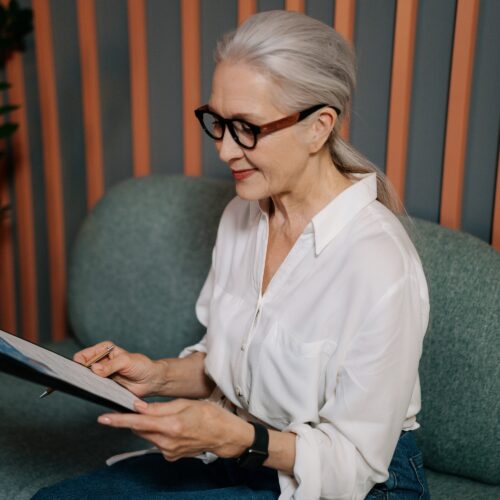 A white, older woman with white hair and black, funky glasses is seated on a modern, grey-green sofa. She is looking at a resume on a clipboard in her hands. She is wearing a white, collared shirt and black trousers. Behind her, there are thin vertical stripes of wood panelling between sections painted grey.