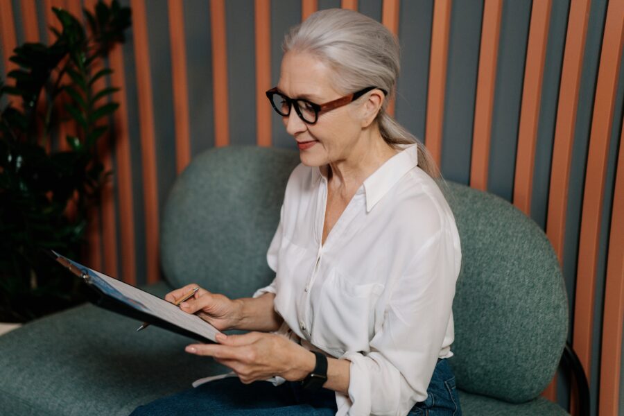 A white, older woman with white hair and black, funky glasses is seated on a modern, grey-green sofa. She is looking at a resume on a clipboard in her hands. She is wearing a white, collared shirt and black trousers. Behind her, there are thin vertical stripes of wood panelling between sections painted grey.