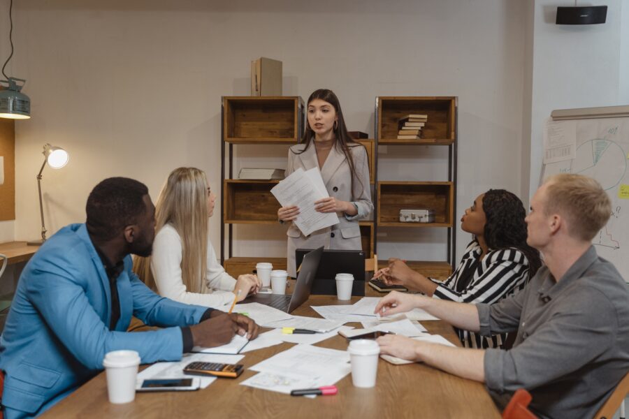 An olive-skinned woman with long, straight brown hair is speaking before her colleagues at a wooden desk. She is standing at the end of the table with papers in her hands; she is wearing a grey, crisp blazer and a dusty coloured turtleneck sweater underneath. Her coworkers are looking at her as she speaks, and they have coffee cups, papers, and other office supplies on the table at which they are seated.