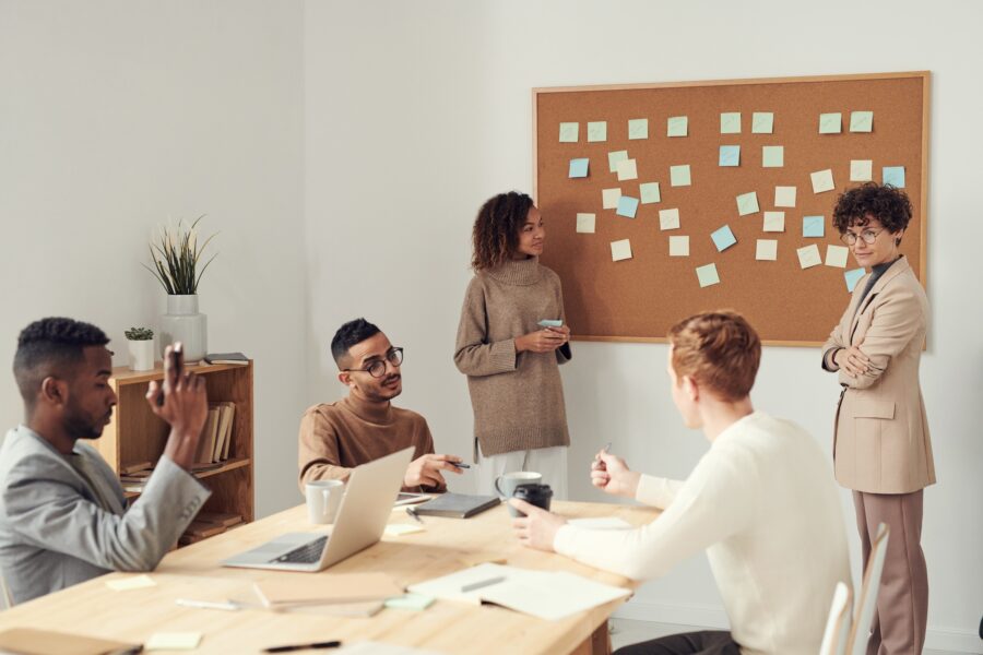 Five young workers are sitting and standing around a light wood table in a modern office setting. The workers are diverse in gender and race, but are all wearing Earth-toned and neutral clothing, such as fine knitted sweaters and lightweight suits in their office setting. They are all looking at their laptops, each other, or the cork board on the far wall that has many sticky notes attached to it.