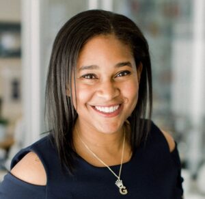 A headshot of Belinda Aramide from the shoulders up. She is a Black woman with straight, dark brown hair parted on the side, bright smiling eyes, and a confident smile. She is wearing a dark navy formal dress or fancy top with a cold-shoulder cut out and a silver necklace with the letter G on it. 