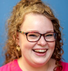 A headshot of Bethany Goldson. She is a white woman with copper-coloured red hair styled in a half-up, half-down ponytail. Her hair has tight curls. She is wearing dark, rectangular glasses, a bright pink shirt, and is smiling openly at the camera, her eyes reflecting her large smile. 
