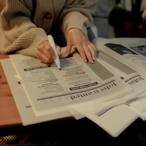 A person's hands are shown placed on top of a newspaper opened up to the Jobs Wanted page. The person has a red marker in their hand and is circling a job posting in the newspaper.