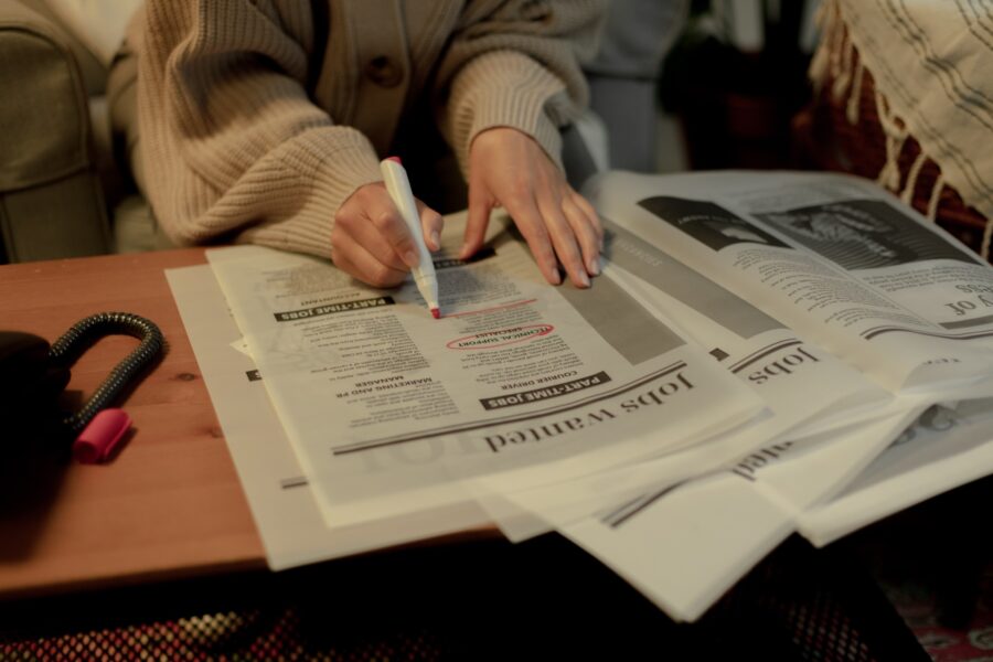 A person's hands are shown placed on top of a newspaper opened up to the Jobs Wanted page. The person has a red marker in their hand and is circling a job posting in the newspaper.