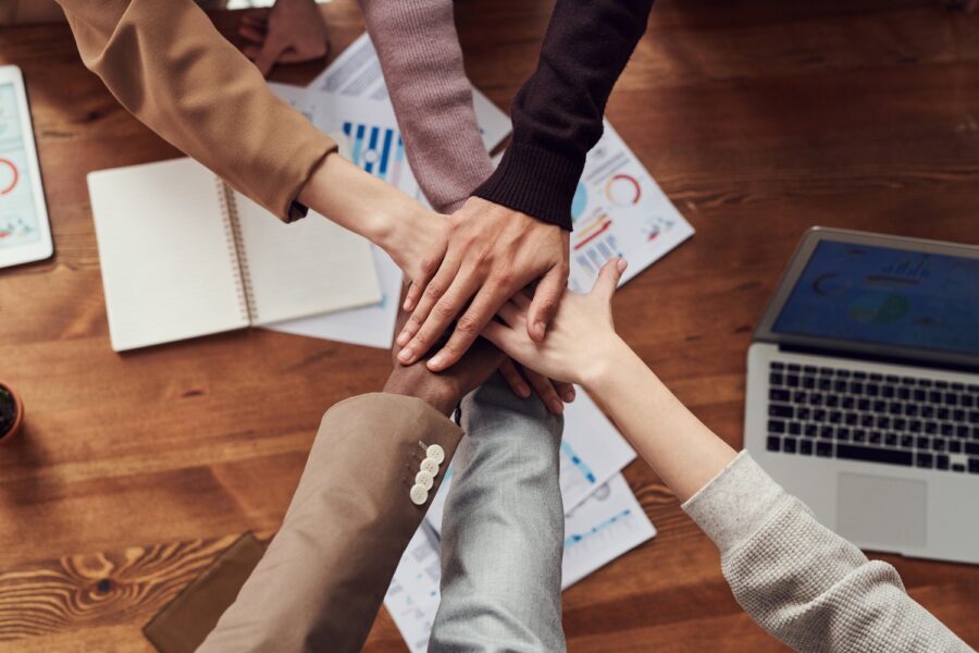 Six people of various skin tones have their hands in the middle of a dark wooden table to show they are part of a team. They are each wearing either a blazer of a neutral, Earth-toned colour, or a lightweight knitted sweater. On the table below their hands are notebooks and pieces of paper with graphs and charts on them. There is a MacBook laptop in the lower righthand corner.