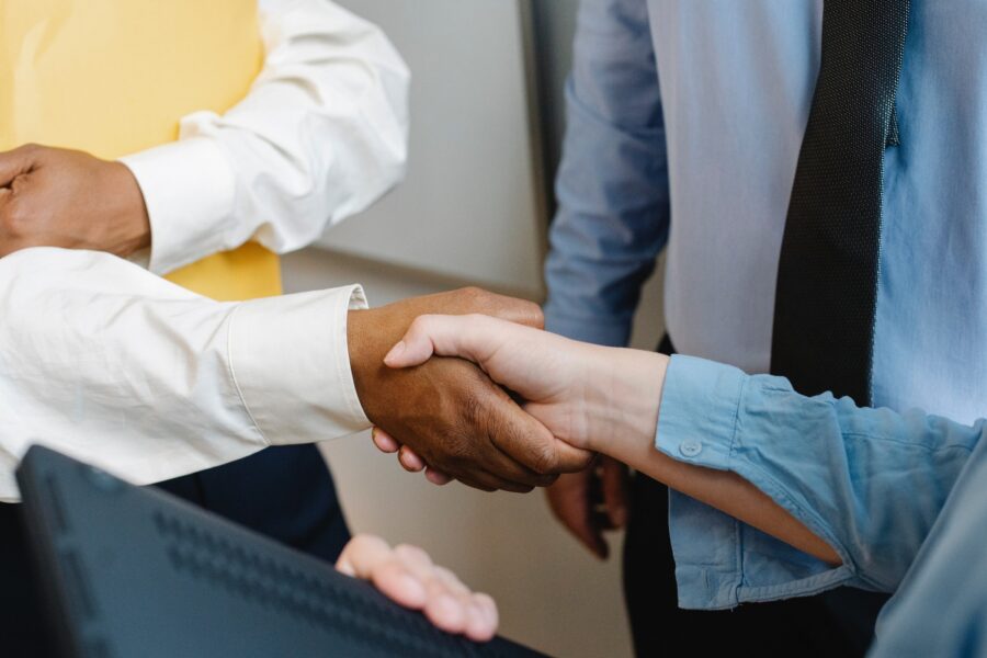 Three people in business attire are standing in a triangular formation facing each other. Two of the people are shaking hands. Only their arms and shirts are visible in the photo.