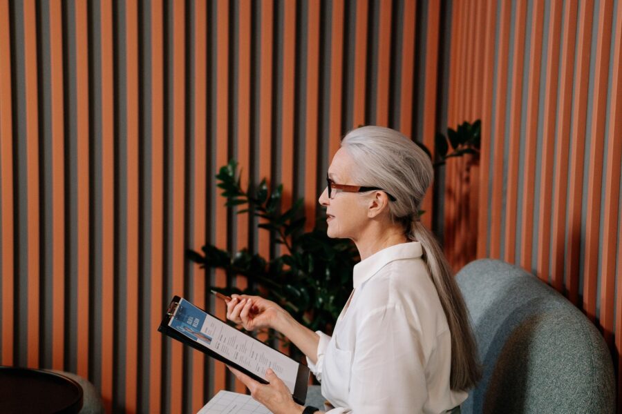 A white woman with long, straight, white/grey hair is sitting on a modern, grey coloured sofa in a funky office space. The walls are panelled with thin wood trim and there is a large plant in the corner. The woman is wearing jeans, a white blouse, glasses, yellow leather loafers, and is holding a clipboard with a candidate's resume on it.