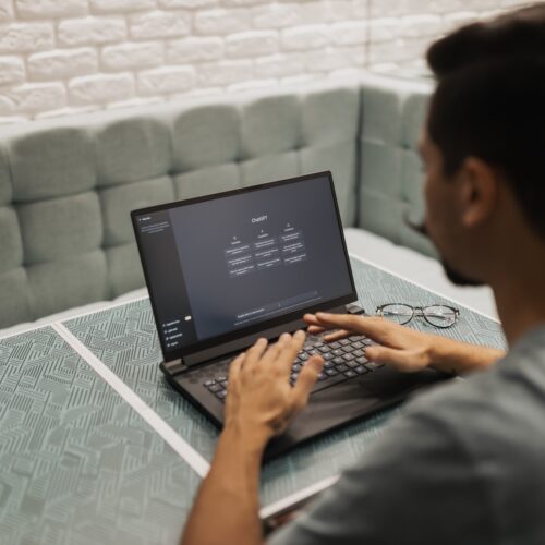 A person with brown skin and dark hair wearing a grey t-shirt is sitting at a grey, modern table with fabric benches on either side. The person is typing on their black laptop, which has the main page to Open AI on the screen.
