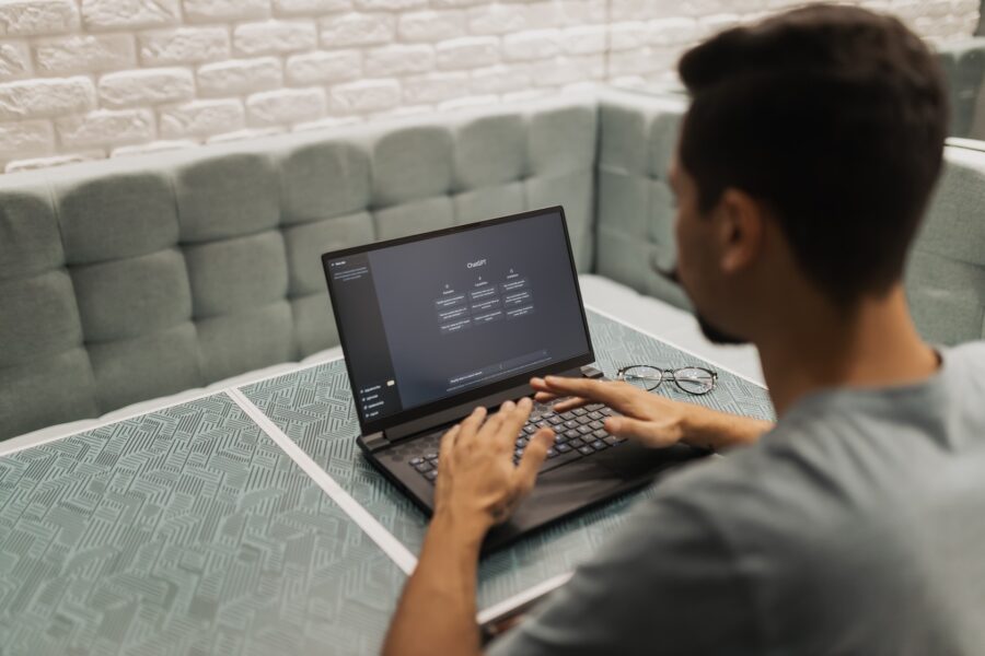 A person with brown skin and dark hair wearing a grey t-shirt is sitting at a grey, modern table with fabric benches on either side. The person is typing on their black laptop, which has the main page to Open AI on the screen.