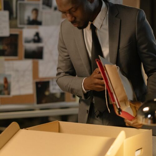 A business man is standing in an office space that is dark save for a few lamps turned on. He is looking into a cardboard box on a table, holding documents and folders under his arm, as if he is about to pack them away. He is dressed in a grey wool suit with a button-up white shirt and a dark tie.