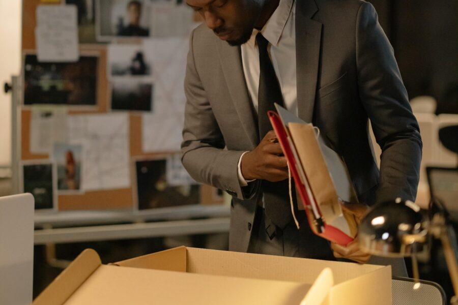 A business man is standing in an office space that is dark save for a few lamps turned on. He is looking into a cardboard box on a table, holding documents and folders under his arm, as if he is about to pack them away. He is dressed in a grey wool suit with a button-up white shirt and a dark tie.