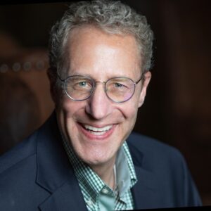 A headshot of Gary Zermuehlen. He is a man in his 50-60s with fair skin, greying hair, and round glasses. In his headshot, he is smiling openly at the camera, wearing a navy suit and blue gingham button up shirt.