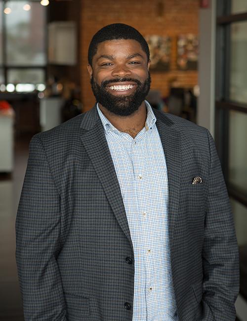 Kaleem Clarkson is a Black man with a short, tidy beard and short cut hair. In his headshot, he is wearing a grey woven suit with a blue button-up shirt underneath and has a large, friendly smile. Behind him, there is a modern-looking office space with a red brick wall, hanging lights, and sleek black architectural features. 