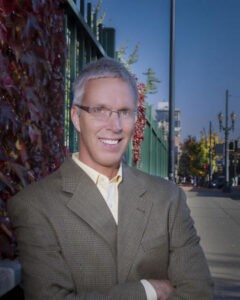 Jeff Schneider is a white man with light grey, short hair, metal glasses, and light coloured eyes. In his headshot, he is in front of a city street with a wall behind his left shoulder and a sidewalk behind the other. He is wearing a grey suit and a light button up shirt, and has his arms crossed over his chest. He is smiling gently while looking at the camera. 