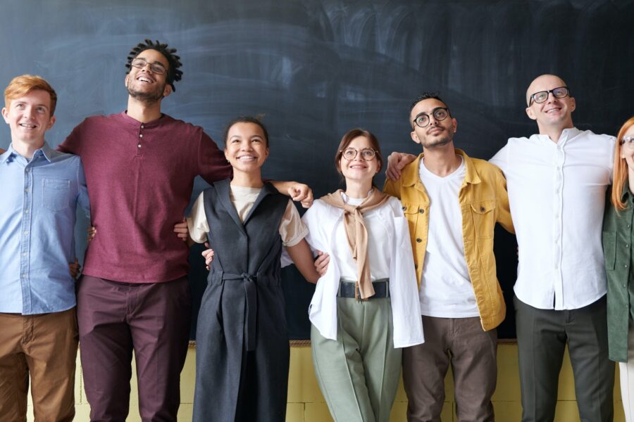 A group of 7 employees are lined up in front of a chalkboard with their arms around each other's shoulders. They are all looking above the camera, smiling, and are all dressed in casual button-down shirts, trousers, and knit shirts in neutral and warm tones, like mustard and maroon. The employees are all a variety of races, genders, and heights, but are mostly young.