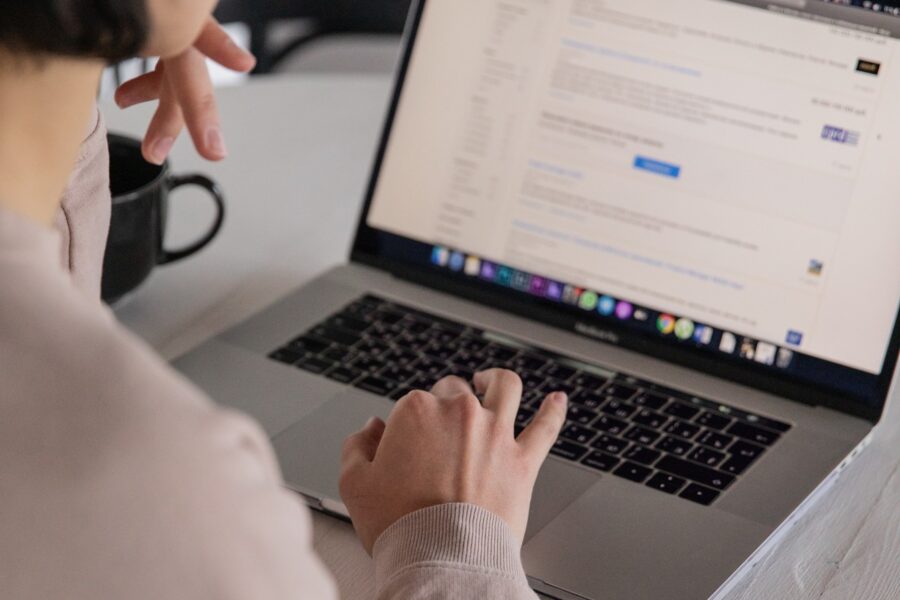 A white woman is sitting at a white desk with one hand on the trackpad of a silver laptop and the other hand at her chin contemplating something she is reading on the screen. Her screen is blurred, but there is black text on a white screen.