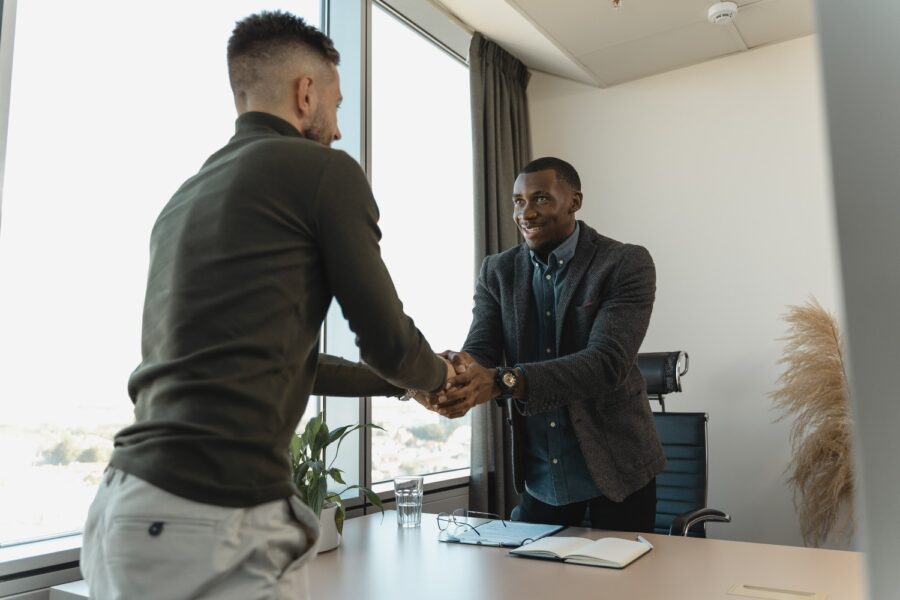 Two men are standing, leaning over a light wooden table in a modern office space. They are both dressed in business casual dress--one in trousers in a turtleneck, the other in casual trousers and a sports jacket with a button-up shirt and tie. They are both smiling and looking at each other.