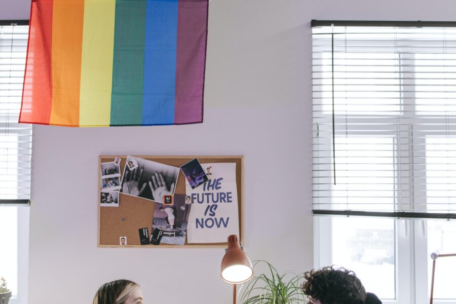 Two workers are sitting at a white desk in a minimalist office space. Above them is a rainbow flag, a lamp, a plant, and a cork board with some images posted to it. Notably, one says "The Future is Now".