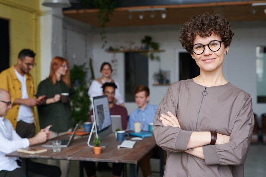 A white-presenting woman with short, brown hair is posing with her arms crossed in the foreground of a modern office space with a sleek table and plants hung on the wall behind her. Around the table are six other workers, all of whom are in button-up shirts and are holding mugs or mobile phones, or are looking at a laptop on the table. The woman in the foreground is wearing black, round glasses and a sleek, taupe-coloured jacket, and is smiling with her arms crossed across her chest.