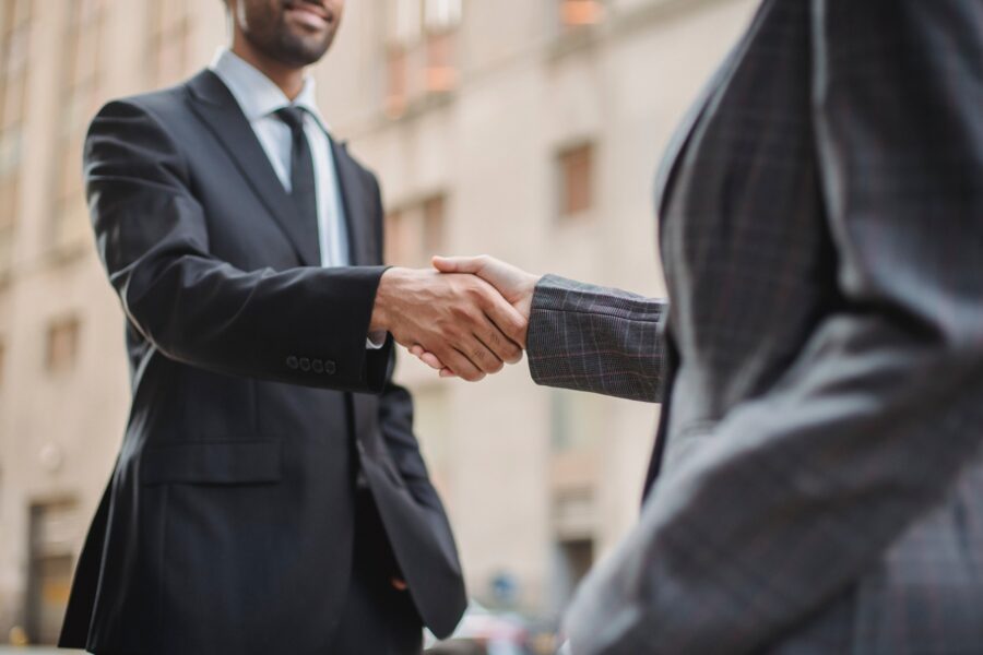 Two people in suits are shaking hands outside an office building. The one in the background, who is more in shot than the other, is a man with light brown skin with a shortly trimmed brown beard. He has a calm smile on his face. The one in the foreground only has their torso in view of the camera.