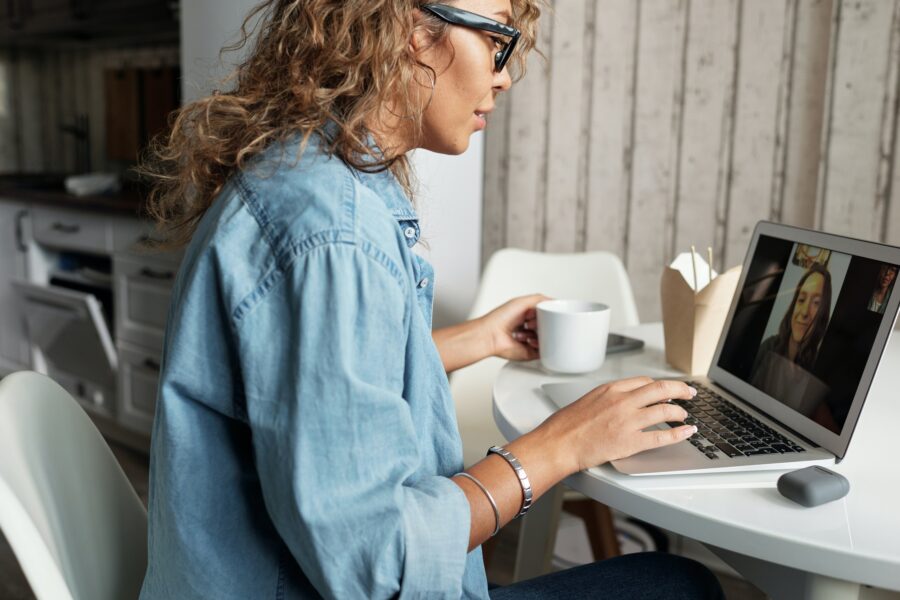 A woman with light brown skin and light brown hair is sitting at a round white desk in her home. She is facing her laptop, which is open on a Zoom or Microsoft teams call with a coworker. In her hand is a white mug; there is also leftover take out food on the table beside her.