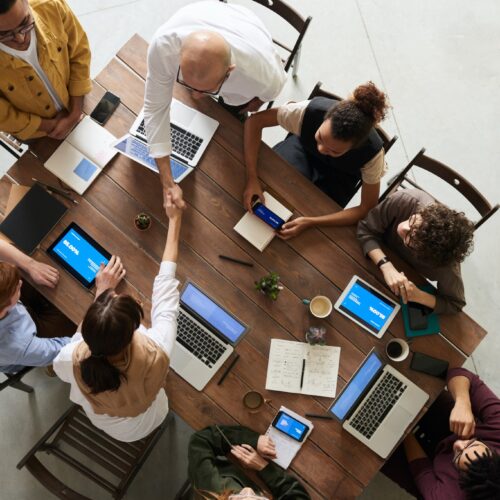 A Birdseye view of a desk shows eight workers sitting on black wooden folding chairs. They are all on laptops or tablets and all have the same blue screen pulled up. Two workers are reaching across the table to shake hands with one another. The workers range in age, race, and gender from what is visible from the Birdseye view: a bald man, a woman with Black, curly hair, a man with red, short hair, and young women with longer, brown hair are present at the table. They are all dressed in neutral business casual clothing, including button-up shirts and cardigans or sweater vests.