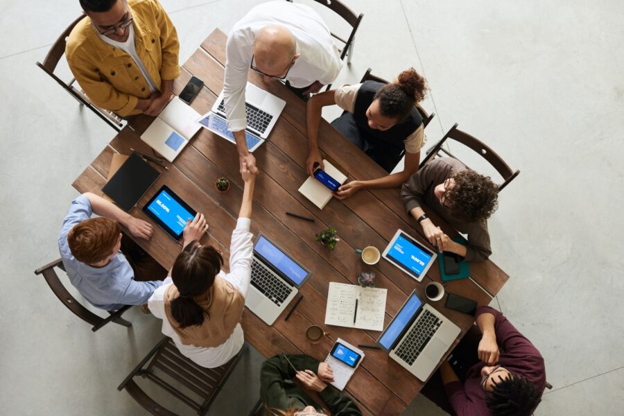 A Birdseye view of a desk shows eight workers sitting on black wooden folding chairs. They are all on laptops or tablets and all have the same blue screen pulled up. Two workers are reaching across the table to shake hands with one another. The workers range in age, race, and gender from what is visible from the Birdseye view: a bald man, a woman with Black, curly hair, a man with red, short hair, and young women with longer, brown hair are present at the table. They are all dressed in neutral business casual clothing, including button-up shirts and cardigans or sweater vests.