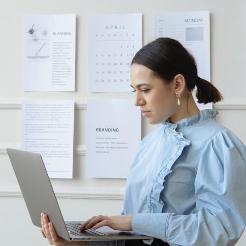 A white woman is standing in a white office space with a silver laptop in one hand. She is typing on the keyboard with the other, and is looking contemplatively at the screen. She has her dark brown hair slicked back into a short, low ponytail and has a ruffled powder blue blouse on. Behind her, there are pieces of paper with text on it taped to the wall.