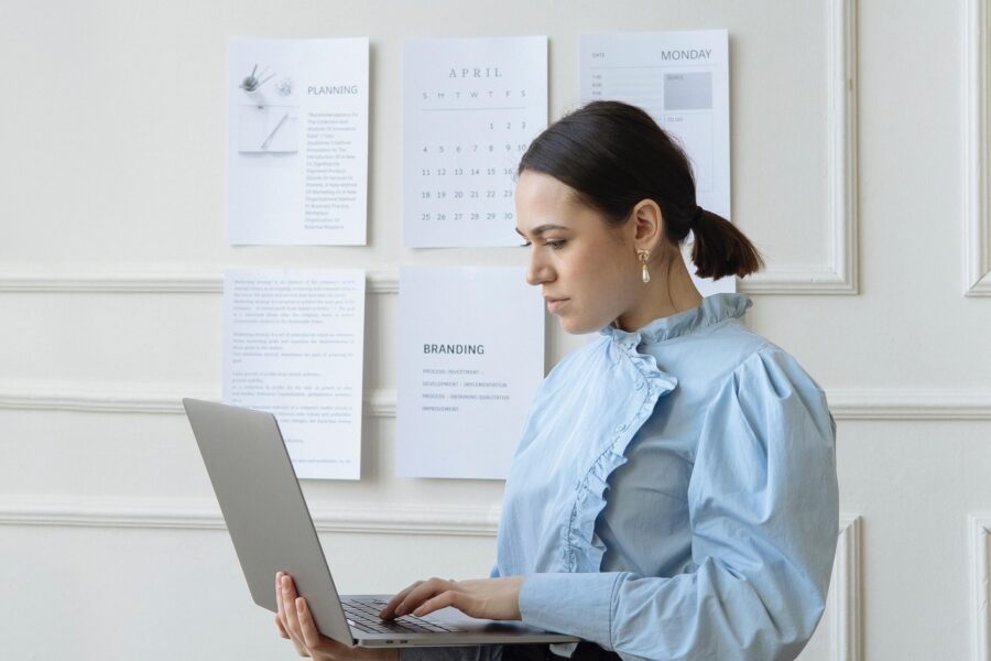 A white woman is standing in a white office space with a silver laptop in one hand. She is typing on the keyboard with the other, and is looking contemplatively at the screen. She has her dark brown hair slicked back into a short, low ponytail and has a ruffled powder blue blouse on. Behind her, there are pieces of paper with text on it taped to the wall.