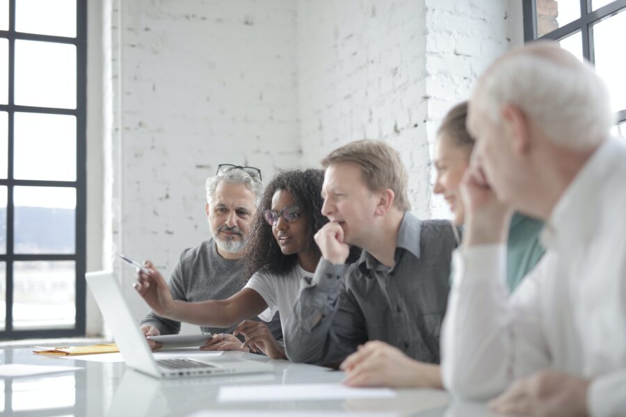 A group of workers are all sitting on the same side of one long desk looking at a white laptop in front of them. One of them, a Black woman, is sitting forward, leaning into the laptop to point at the screen with her pen. The other workers around her are looking at where she is pointing and are listening attentively.