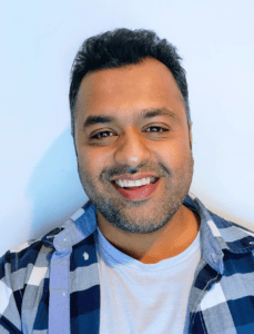 Sam Doshi is a man with dark hair styled upwards off of his face. His headshot shows him in front of a white wall in a plaid shirt layered on top of a plain t-shirt. He is smiling warmly at the camera, showing his straight teeth. 