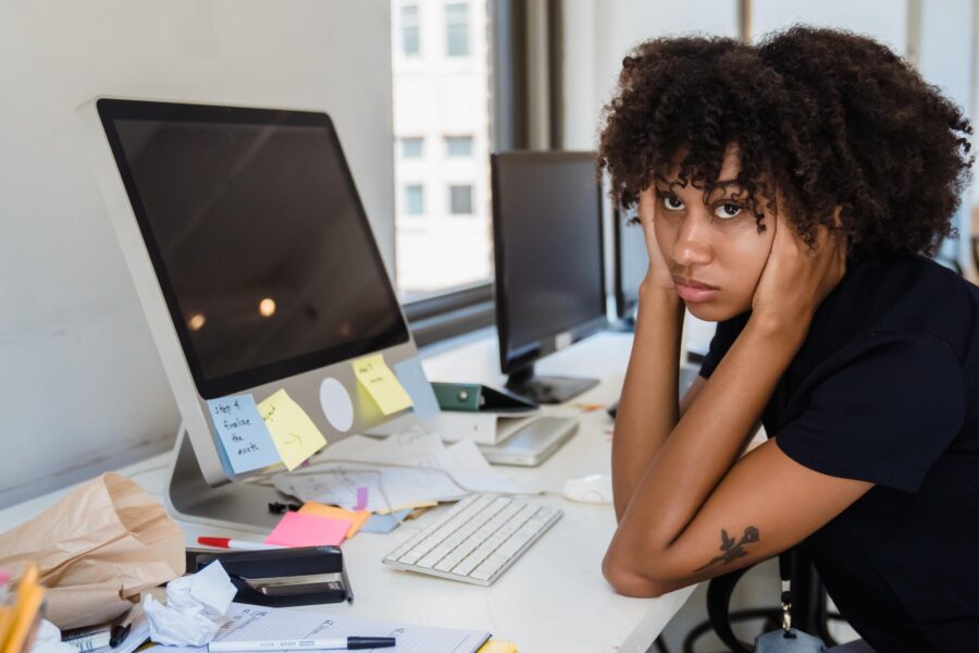 A Black woman is sitting at a long, thin desk by an office window. On the desk are multiple desktop monitors and keyboards, along with pens, papers, and other office supplies. She is looking just above the camera with her hands on her face, looking bored and frustrated.