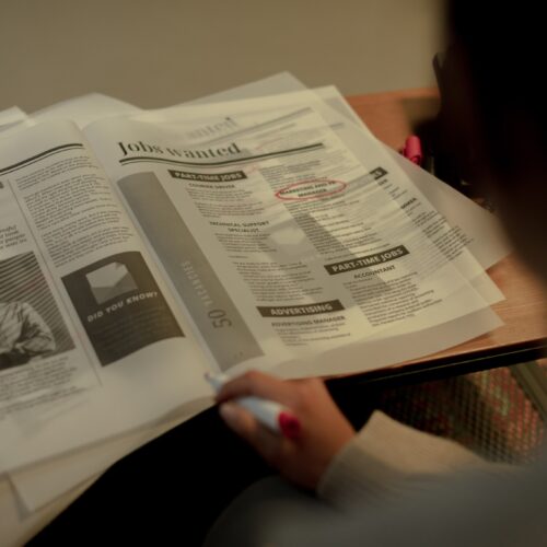 A newspaper is laid open on a wooden table, and the camera is pointed toward it from over a person's shoulder. The person is out of focus, but the newspaper is in focus and reads "Jobs Wanted" on the page open. One job posting is circled in bright red marker.