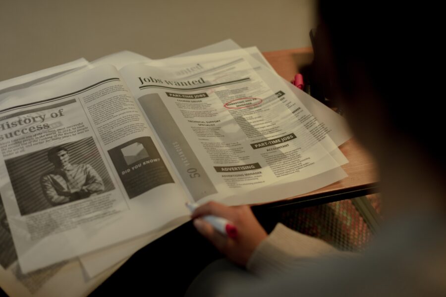 A newspaper is laid open on a wooden table, and the camera is pointed toward it from over a person's shoulder. The person is out of focus, but the newspaper is in focus and reads "Jobs Wanted" on the page open. One job posting is circled in bright red marker.