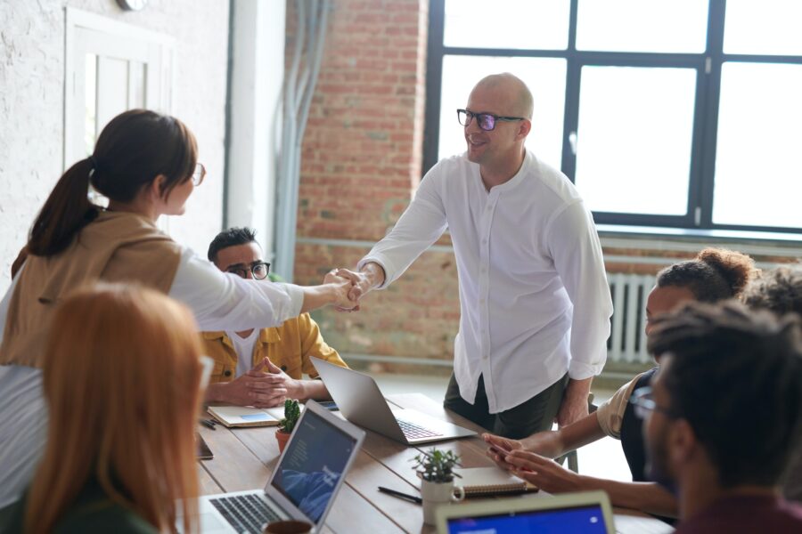 A team of office workers is sitting around a large desk in a room with large windows and exposed brick walls. Two people are standing at the table, reaching over the table to shake each other's hand. On the table, there are also laptops, notebooks, and pens.