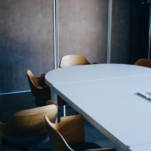 In an otherwise empty board room, there are modern, wood chairs turned outwards to show people were once sitting and them and perhaps left abruptly without tucking them back in. The space is modern with dark, charcoal painted walls and a sleek white conference table.