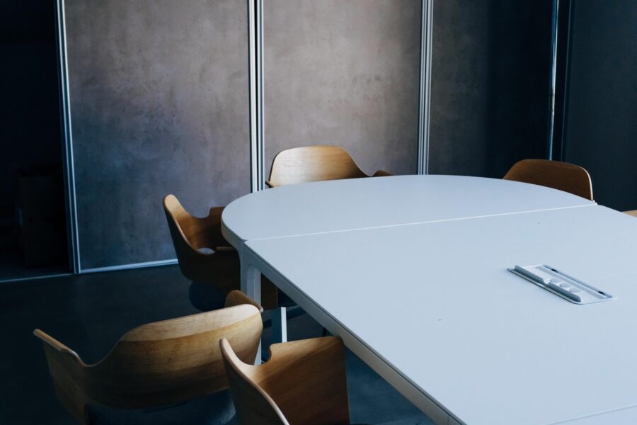 In an otherwise empty board room, there are modern, wood chairs turned outwards to show people were once sitting and them and perhaps left abruptly without tucking them back in. The space is modern with dark, charcoal painted walls and a sleek white conference table.